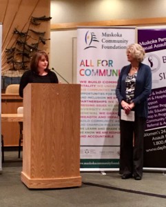 A woman stands at a podium, speaking, while another woman looks on. A banner behind the women reads: Muskoka Community Foundation, All for Community.