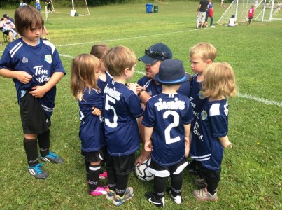 A coach huddles in conversation with the players of his children's soccer team on the sidelines of a soccer pitch.
