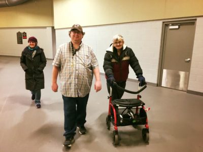 Three adults, one with a walker, walk on an indoor walking track at a recreation centre.
