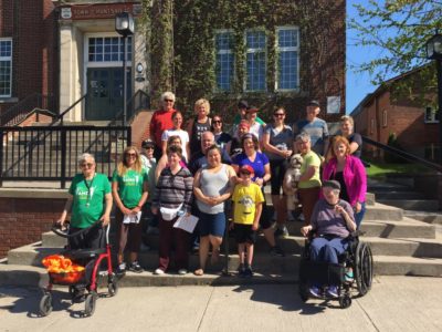 Roughly two dozen people, one with a walker and one sat in a wheelchair, pose for a photo on the steps of downtown Huntsville's town hall.