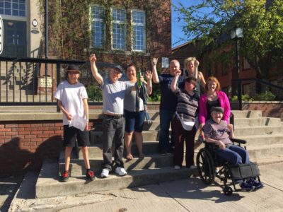 Eight people, including one person sat in a wheelchair, cheer with their hands raised while posing on the steps of downtown Huntsville's town hall.