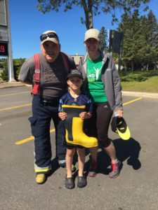 A firefighter poses for a photo with a woman in a Band on the Run T-shirt and a boy holding a yellow and black rubber boot.