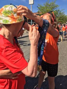 A woman in an orange T-shirt drapes a race finisher medal over the head of a person in a red T-Shirt.