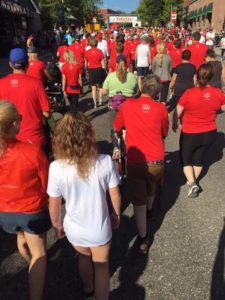 A large group of people in red T-shirts walk toward the start line of a foot race. The photo is taken from the back of the crowd.