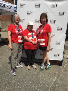 Three people in red T-shirts pose with their race finisher medals in front of a white photo backdrop.