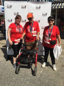 Three people in red T-shirts pose with their race finisher medals in front of a white photo backdrop.