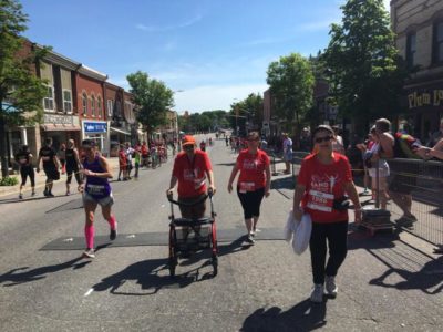 Four people run or walk to the finish line of a foot race while spectators cheer on the sidelines.