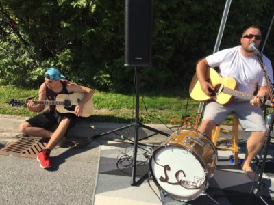 A man sitting on a roadside curb plays a guitar, while another man beside him sits on a stool playing a guitar and kick drum.