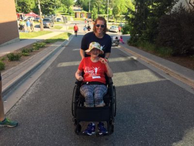 A person wearing a red Band on the Run T-shirt is sat in a wheelchair. A support person assists them by pushing the chair.