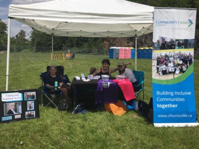 Three people sit under an opened sided tent in a park. They have a table with information and merchandise in front of them. A banner beside them reads: Community Living Huntsville, Building Inclusive Communities Together