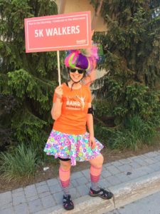 A woman in an orange T-shirt, floral print skirt, high socks, running shoes and colourful bike helmet holds a sign that reads: 5K Walkers.