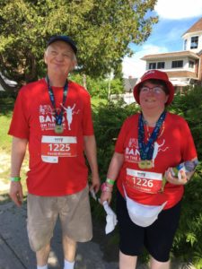 A man and a woman in red Band on the Run T-shirts smile for the camera. They have race finisher medals draped around their necks.