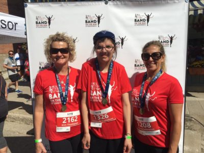 Three women in red Band on the Run T-shirts pose for the camera in front of a white photo backdrop with their race finisher medals.