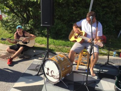 A man sitting on a roadside curb plays a guitar, while another man beside him sits on a stool playing a guitar and kick drum.