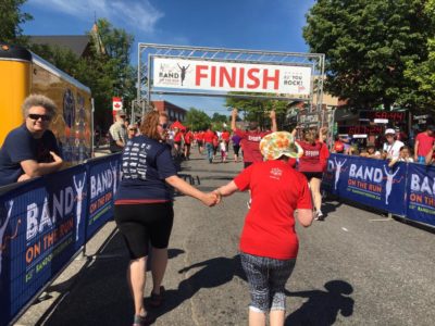 Two people run toward the race finish line while holding hands. One of the women, who is slightly ahead, looks back in encouragement for the other.