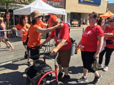 A woman in an orange T-shirt drapes a finisher medal over the shoulders of a man in a red T-shirt. He pushes a mobility walker.