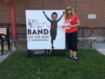A person poses for a photo by positioning their face in a hole cut out of a large race logo poster. A woman in a red Band on the Run T-shirt stands beside them and smiles.