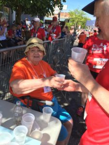 A woman in an orange T-shirt is seated. She is handing a beverage in a plastic cup to a race participant in a red T-shirt, who is accepting the cup. He is also holding a second cup.