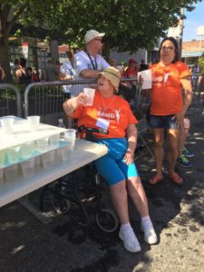 A woman in an orange T-shirt, seated, readies to hand beverages in plastic cups to passing runners.