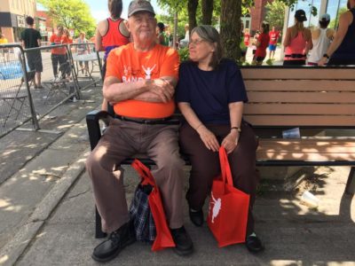 A man in an orange Band on the Run T-shirt sits on a bench near the race route. A woman sits close beside him, smiling while she looks at him.