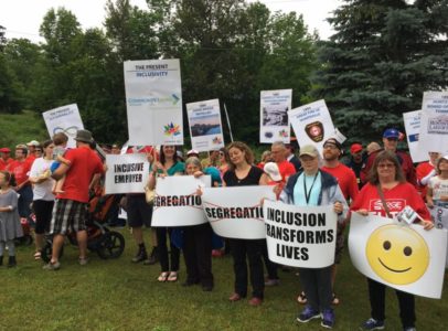 A large group of smiling people holding signs toward the camera. One sign reads: Inclusion Transforms Lives. Another sign has the word "segregation" crossed out in red ink