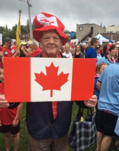 A man in a tall red-and-white faux-felt hat holds a Canada flag while posing for the camera.