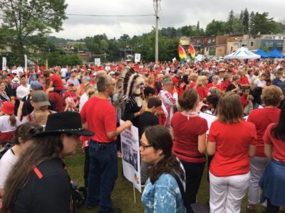 Hundreds of people in red and white gathered in a downtown riverfront park. The photo is taken from the side of the crowd.