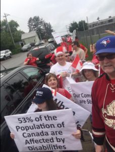 Roughly a dozen people wearing red and white walk by the camera. One person is holding a sign that reads: 53 per cent of the population of Canada are affected by disabilities.
