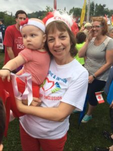 A woman with a red and white Canada hat and a white T-shirt that reads Peace Love and Inclusion. She holds an infant also wearing red and white.