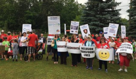 A large group of smiling people holding signs toward the camera. One sign reads: Inclusion Transforms Lives. Another sign has the word "segregation" crossed out in red ink