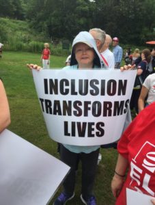 A woman in a rain coat holds a sign that reads: Inclusion Transforms Lives