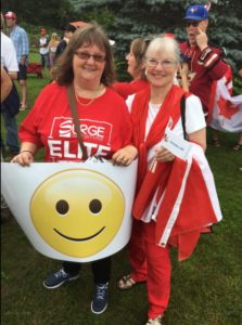 Two women in red and white hold a sign with a big yellow happy face illustration on it.