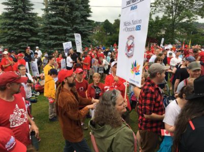 A large group of dozens of people in a park. One holds a sign that reads: Metis Nation of Ontario.