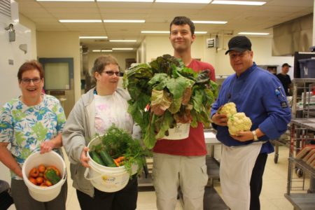 Image shows two women and two men in a commercial kitchen holding armfuls of fresh vegetables while smiling at the camera.