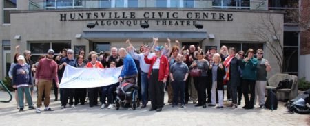 Image shows a large crowd of happy people cheering outside the Huntsville Civic Centre in downtown Huntsville while holding a Community Living Huntsville flag. The flag is white with green and blue text. Text reads: Community Living Huntsville..