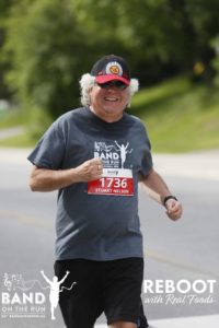 A smiling man in a ball cap running down a paved road. The collar of his T-shirt is damp.