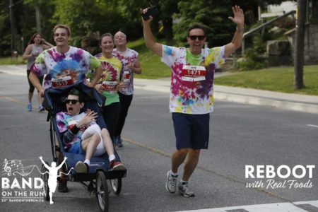 five people in white T-shirts tie-dyed with red, green, blue, purple and yellow jog down a paved residential road. One man is sat in a running stroller, a man pushes the stroller from behind, and another man raises his arms in the air and cheers for the camera.