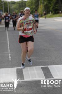 A woman in a tie-dyed T-shirt with a white bandana on her head runs down a paved road. A group of fellow runners is in the distance behind her.