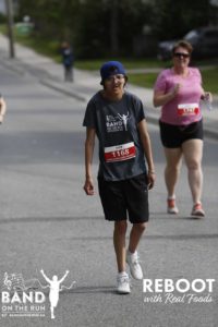 A person in a grey Band on the Run T-Shirt and blue ball cap walks down a paved road. A woman in a pink T-shirt also runs in the background.