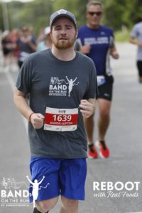 A man in a grey Band on the Run T-shirt, blue shorts and ball cap runs down a paved road. There are more people in the background behind him.