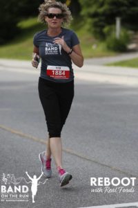 A woman in a grey Band on the Run T-shirt, sport pants and pink-laced running shoes runs down a paved road.