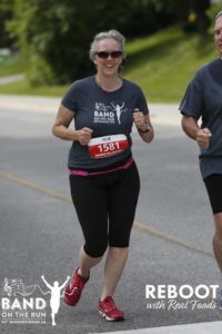 A woman in a grey Band on the Run T-shirt, athletic pants and red running shoes smiles for the camera as she runs passed.