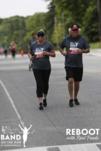 Two people in matching grey Band on the Run T-shirts jog down a paved road.