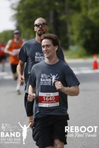 A smiling man with headphones in his ears runs down a paved road. There is a man running behind him in a matching grey T-shirt.