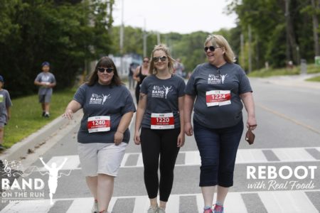 Three women in matching grey T-shirts with race bib numbers pinned to their shirts smile while walking side by side along the race route.