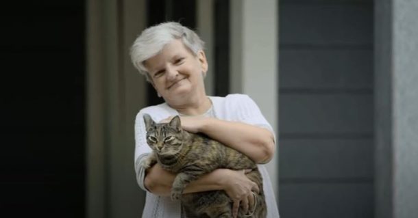 Image shows a smiling woman outdoors holding her pet cat in her arms. She and the cat are both looking at the camera. 