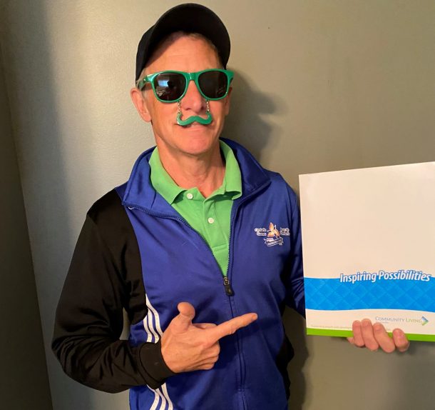 Image shows a man in a green shirt, blue jacket, and green sunglasses with novelty green moustache that dangles over his upper lip. He is holding a folder in one hand and pointing to it with the other. Folder reads: Inspiring possibilities, and includes the Community Living Huntsville logo.