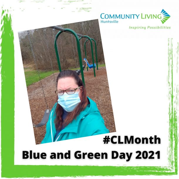 A woman in a green coat wearing a mask outside with a community playground behind her