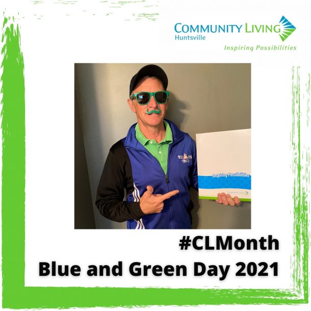 A man in a green shirt, blue running jacket and blue hat dons green sunglasses with a plastic green moustache dangling over his upper lip. He holds a Community Living Huntsville folder that reads "Inspiring Possibilities"