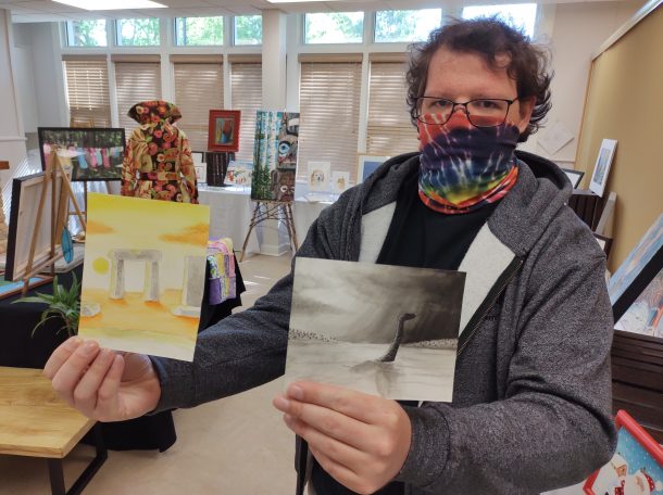 Image shows a man standing in a room filled with art displays. He is wearing a cloth face mask and holding two unframed watercolour paintings. One is yellow and orange in colour, the other is black and white.